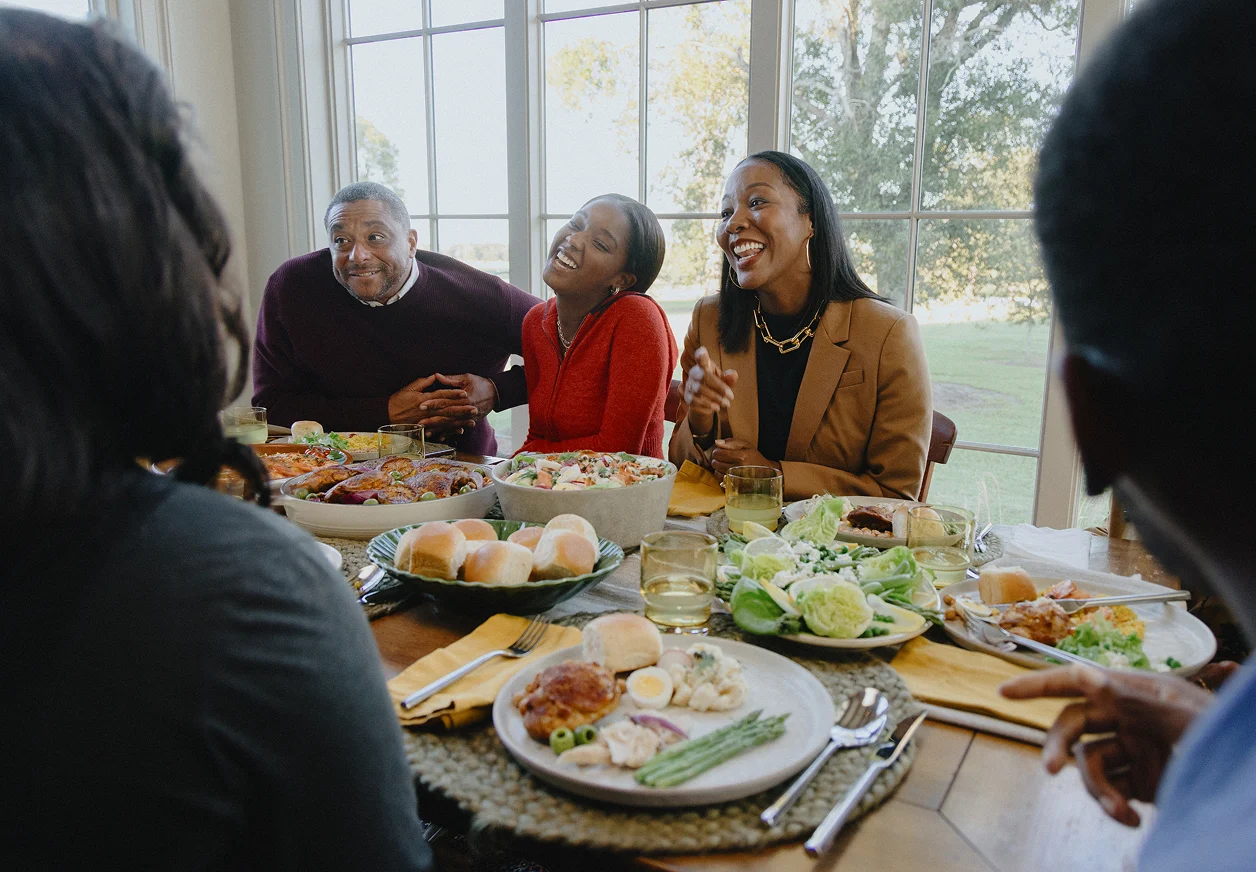 A Black family enjoys happy conversation while gathered around a dining table crowded with dishes of food.