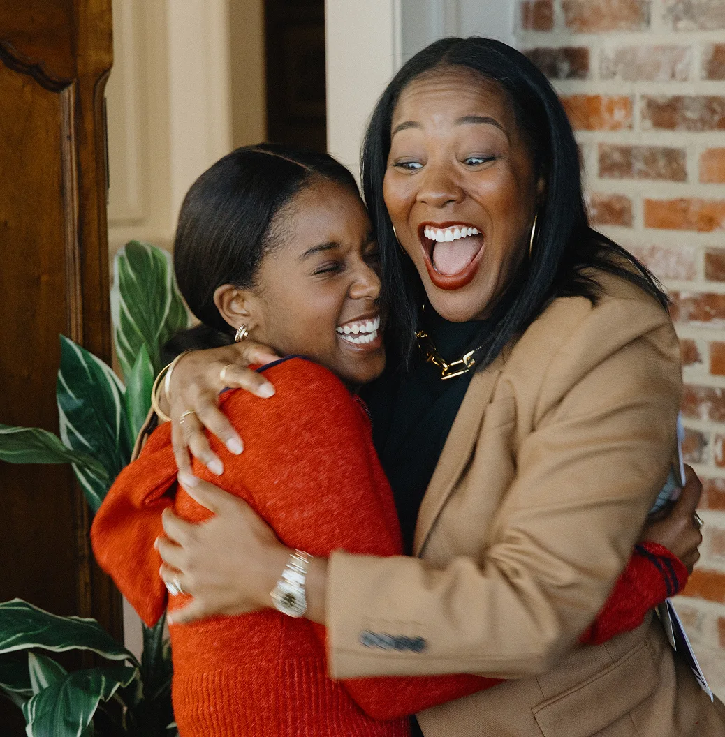 A Black mom in a tan blazer hugs her teenage daughter, sharing  excitement over the new home they just bought with a jumbo loan from Rocket Mortgage.