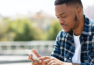 A young Black man wearing a blue plaid flannel shirt researches jumbo loans from Rocket Mortgage on his phone.