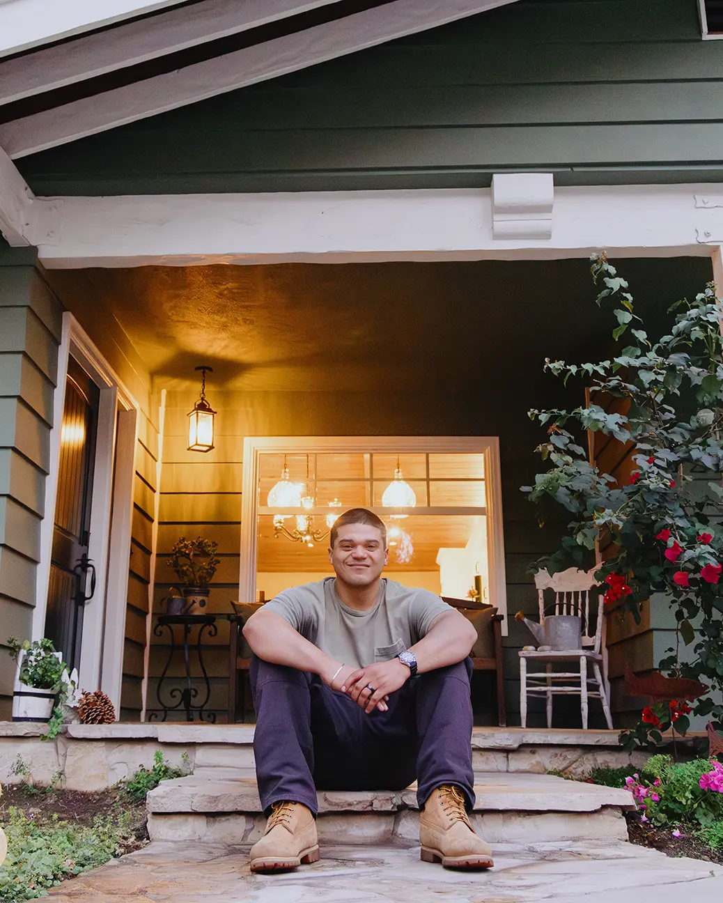 A Hispanic man wearing work boots, brown pants and a tan t-shirt smiles proudly while sitting on the front steps of a stone porch, with a lighted picture window in the background.