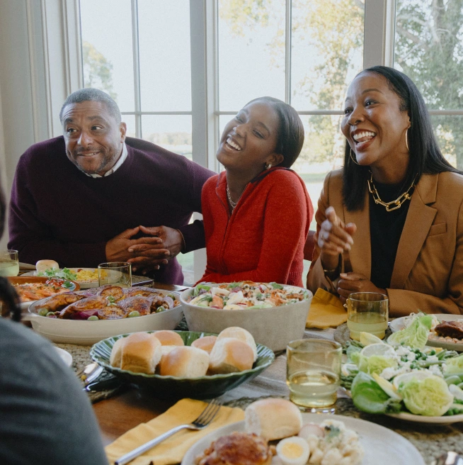 A Black family enjoys happy conversation while gathered around a dining table crowded with dishes of food.